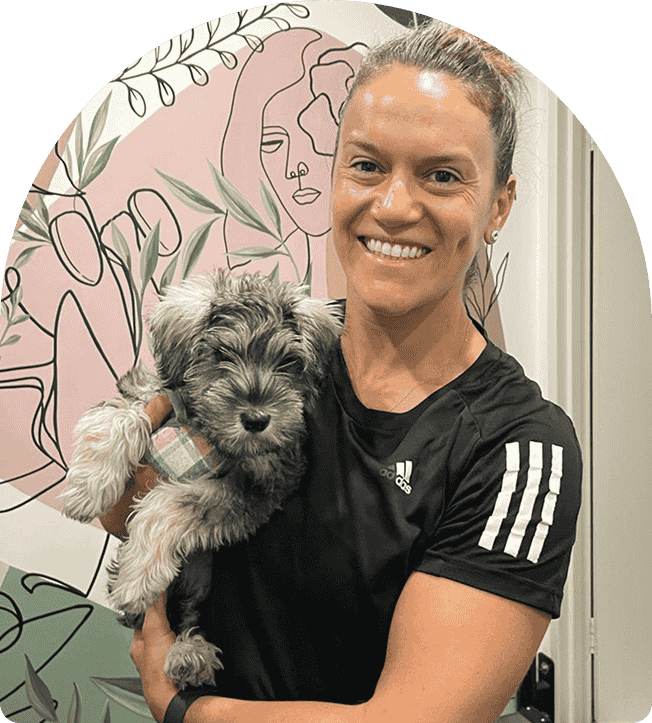 Personal trainer smiling while holding a small dog, set against a colorful, artistic background, representing a supportive and friendly atmosphere for clients at Wellness On Shore in Chiswick.