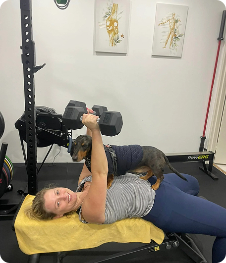 Woman exercising with dumbbells on a gym bench, playful dog on her chest, wellness training environment with fitness equipment and wall art.