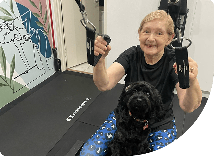 Older woman engaging in fitness training with resistance bands, seated on a gym floor with a black dog beside her, surrounded by a colorful mural, illustrating a supportive and community-focused wellness environment.