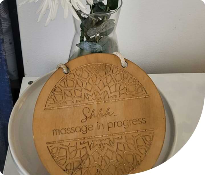Wooden sign reading "Shhh.. massage in progress" with decorative patterns, placed beside a vase with flowers, emphasizing a calming atmosphere for remedial massage therapy at Wellness On Shore.