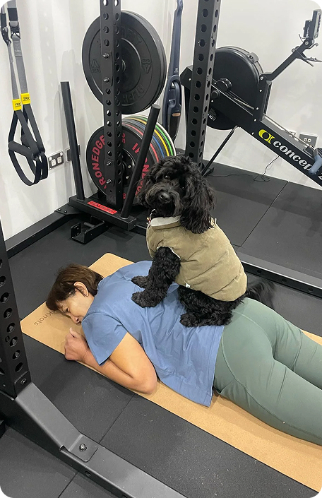 Woman exercising on a mat with a small dog sitting on her back in a gym setting, surrounded by fitness equipment.