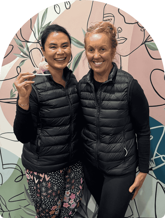 Two smiling yoga instructors wearing black vests, posing together in front of a colorful abstract mural, promoting a welcoming atmosphere for yoga classes at Wellness on Shore.