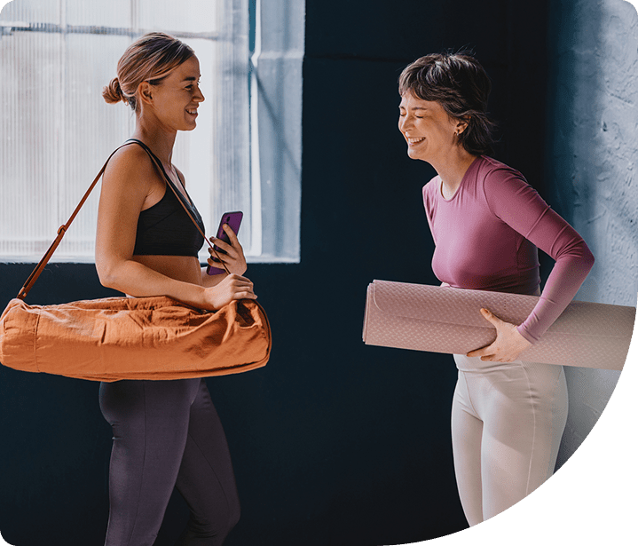 Two women smiling and chatting in a yoga studio, one carrying a yoga mat and the other holding a yoga bag, emphasizing a welcoming atmosphere for beginners at Wellness on Shore.