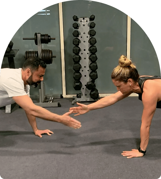 Two individuals engaged in a fitness exercise, performing a push-up high-five, with dumbbells visible in the background, illustrating a supportive and interactive workout environment at Wellness on Shore.