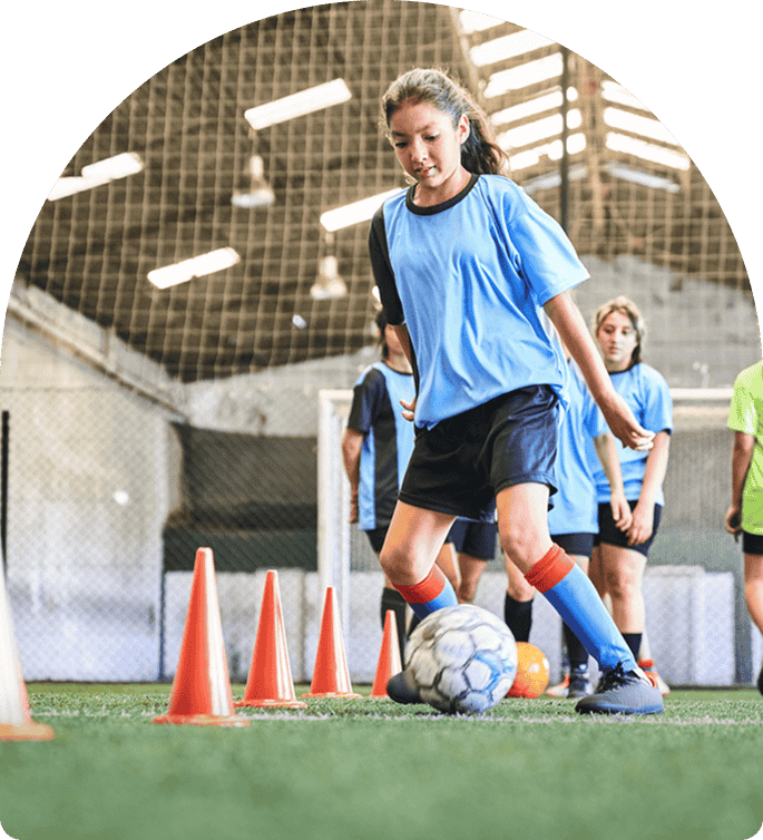 Young female soccer player practicing dribbling skills with a soccer ball and agility cones in a training facility, emphasizing strength and technique for the Girls Soccer Strength Program.