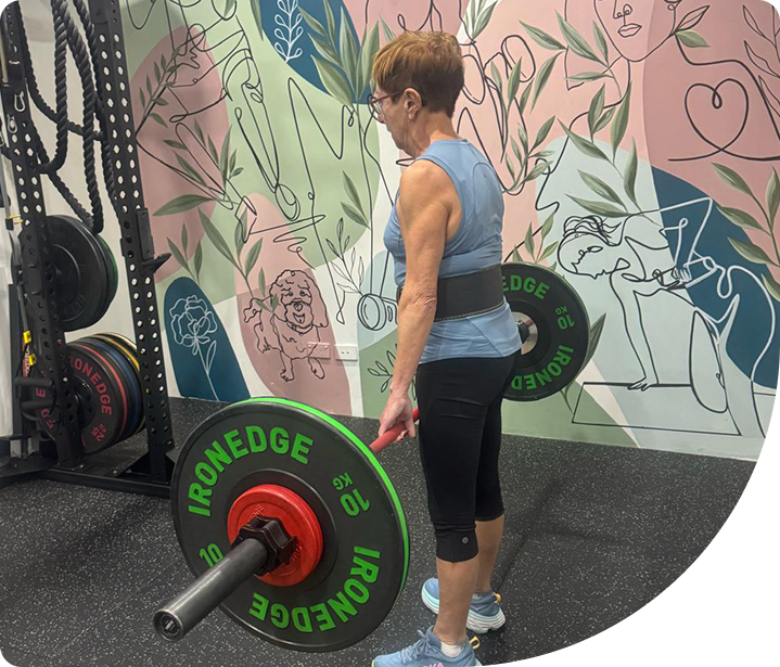 Older woman performing a deadlift with a barbell in a gym, showcasing functional strength training for joint health and mobility, surrounded by colorful wall art.