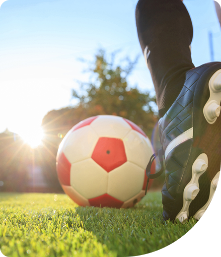 Soccer ball with red and white panels on grass, foot in black cleat positioned to kick, sunlight shining in background, emphasizing youth athleticism and movement.
