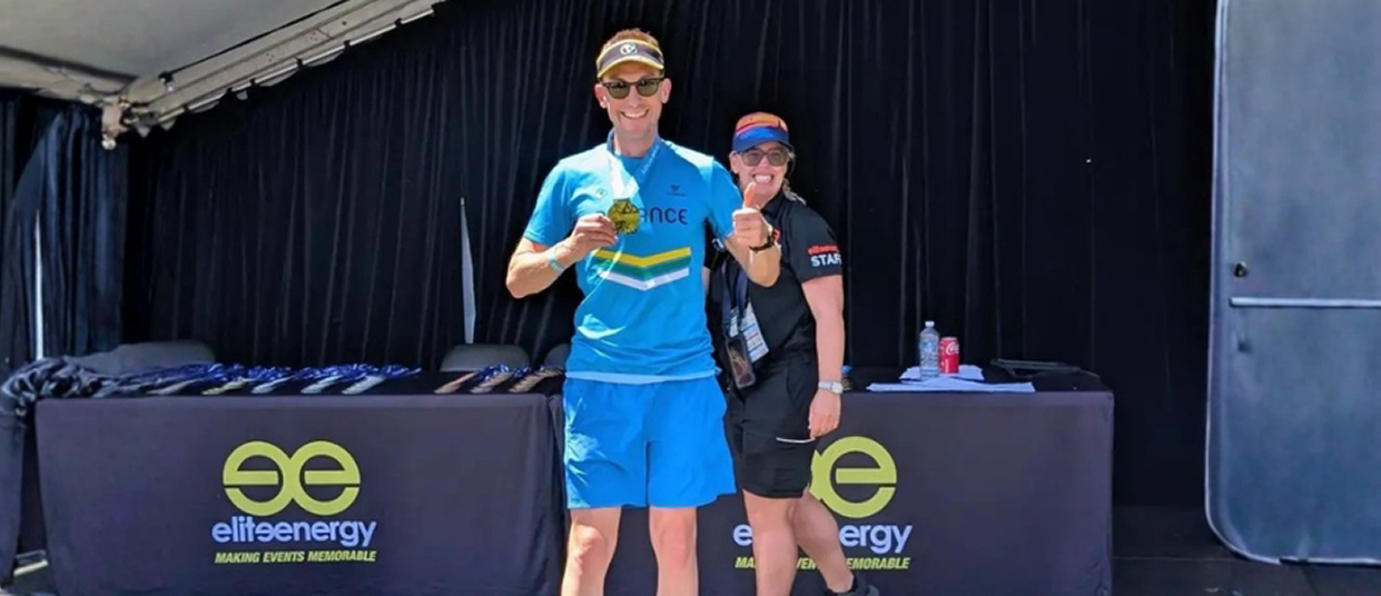 Athlete celebrating with medal at triathlon event, showcasing joy and achievement, with Elite Energy branding in the background.