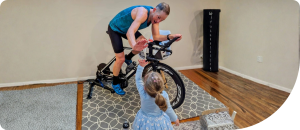 Person cycling indoors on stationary bike, child reaching out, showcasing family fitness and wellness activities.