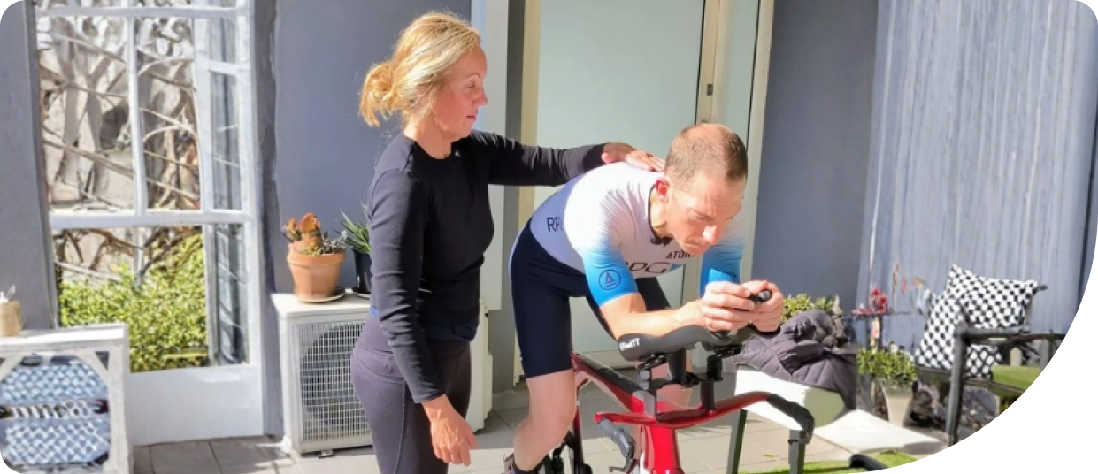 Cyclist receiving guidance on posture and stability while training indoors, focusing on core strength and neuromuscular connection, with a trainer providing support in a bright outdoor setting.