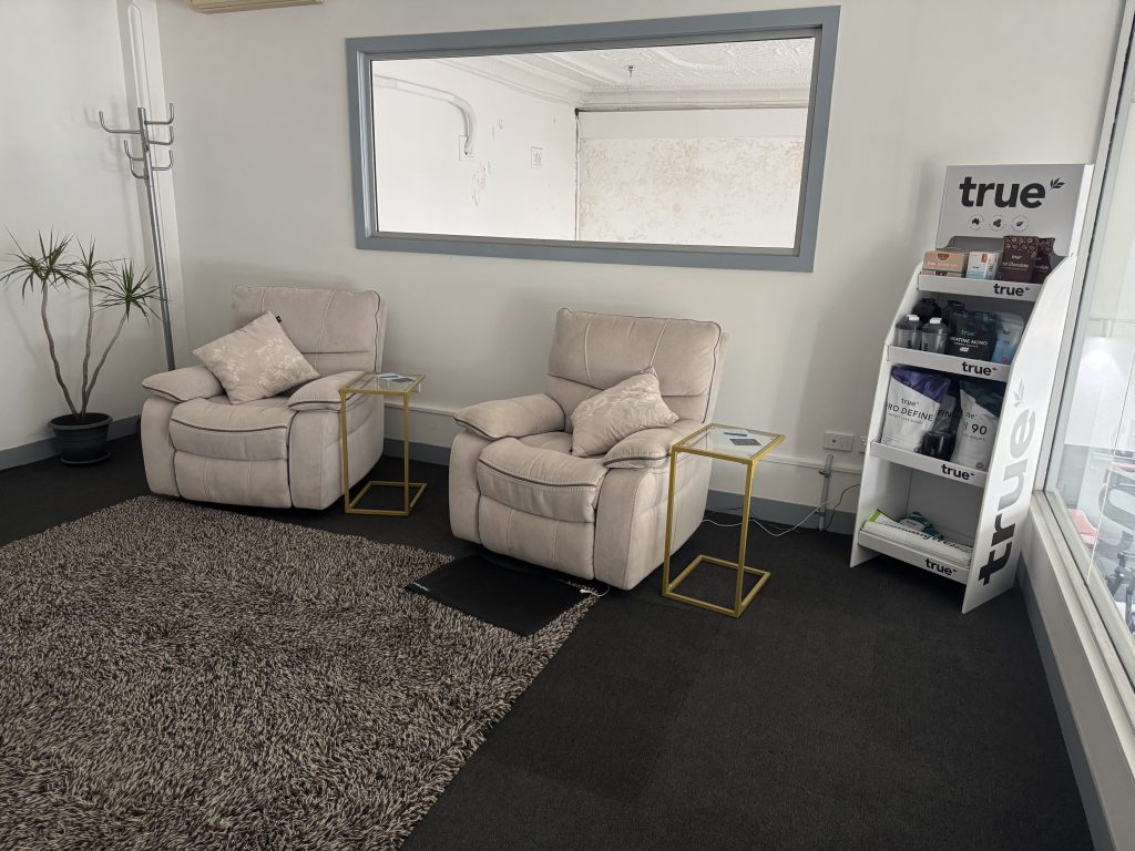 Cozy treatment room at Wellness on Shore featuring two plush armchairs with pillows, a decorative rug, a small side table, and a product display shelf labeled "true."