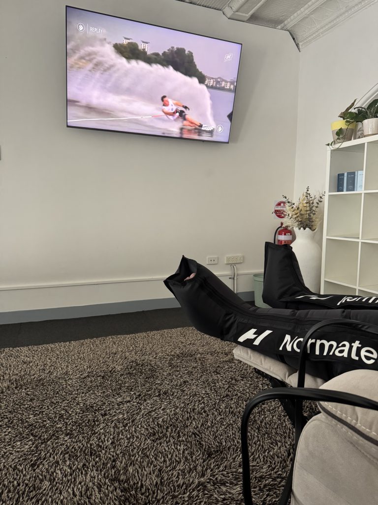 Comfortable wellness treatment room featuring a television displaying water skiing, with Normatec recovery boots on a plush rug, promoting relaxation and health at Wellness on Shore.