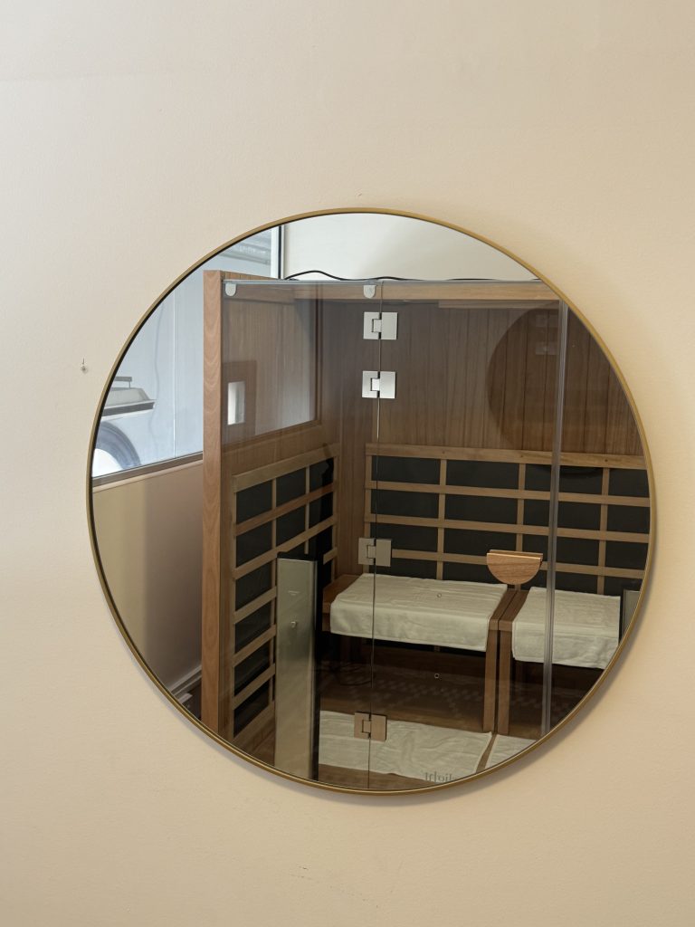 Round mirror reflecting a treatment room at Wellness on Shore, featuring wooden accents, a massage table, and a sauna area, emphasizing a serene wellness environment.