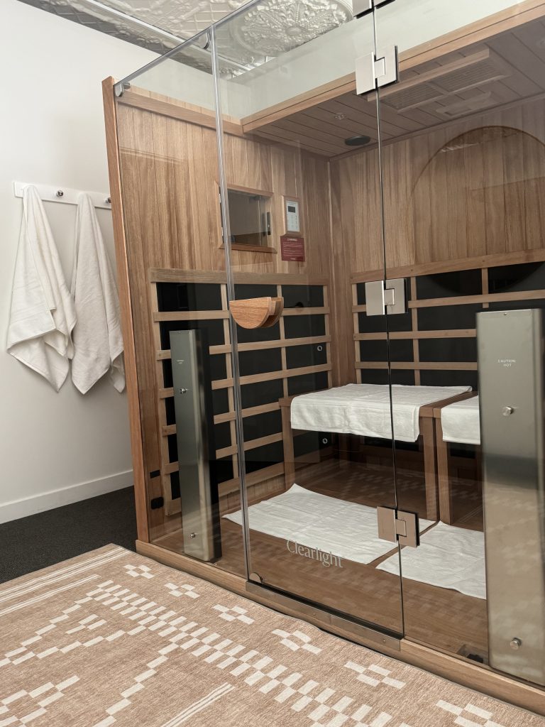 Wellness on Shore treatment room featuring a ClearLight sauna with wooden interior, towels hanging on the wall, and a patterned rug, emphasizing relaxation and wellness services.