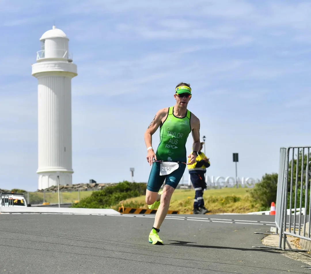 Athlete in green triathlon gear running past a lighthouse during the 2025 Wollongong World Triathlon Championships, showcasing determination and community spirit.