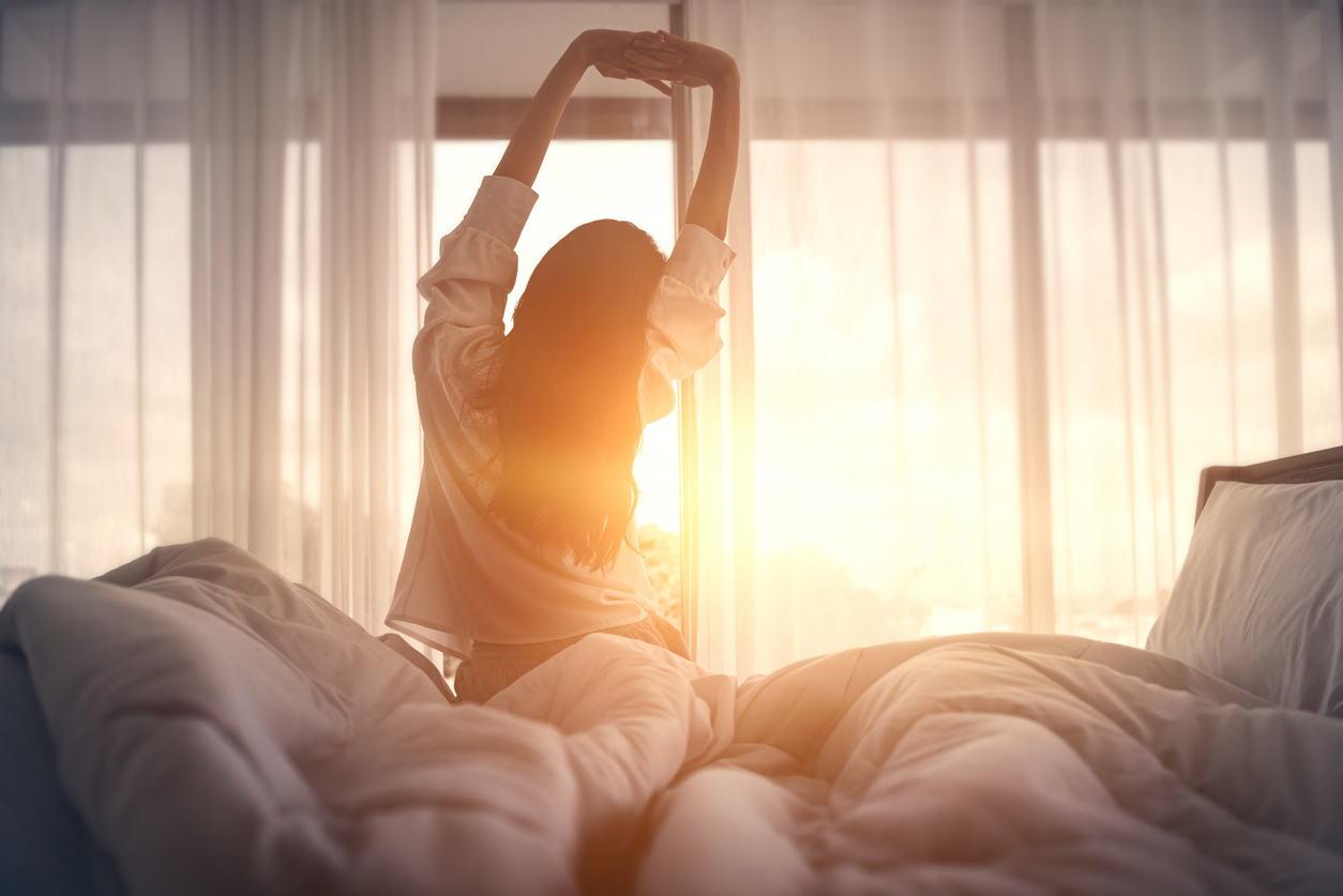 Woman stretching in bed, enjoying morning sunlight, emphasizing the importance of quality sleep for wellness and recovery.