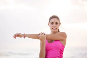 Young woman stretching her arm on the beach, promoting wellness and exercise for recovery and movement education.
