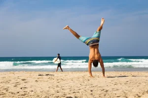 Man performing a handstand on the beach with ocean waves in the background, highlighting active lifestyle and wellness.