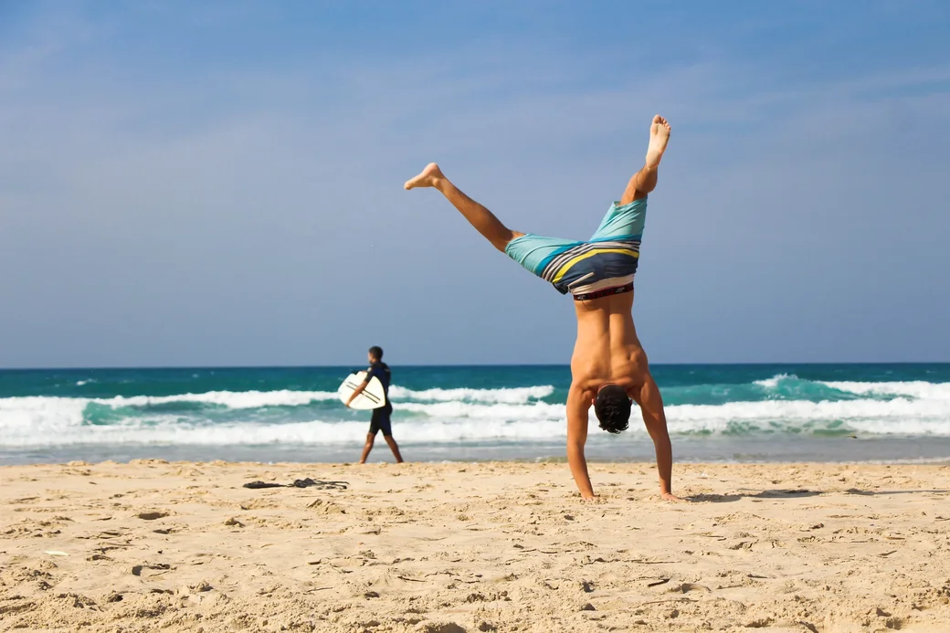 Man performing a handstand on sandy beach with ocean waves in the background, illustrating fun and active lifestyle related to enjoyable exercise.