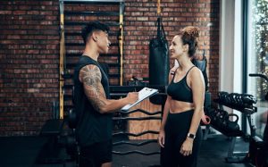 Personal trainer engaging with a client in a gym setting, discussing fitness goals and recovery strategies.
